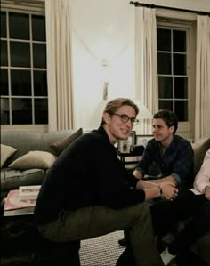 three men sitting on couches in a living room with two windows and one is smiling at the camera