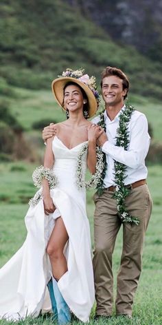 a man and woman standing next to each other in the grass with flowers around their necks