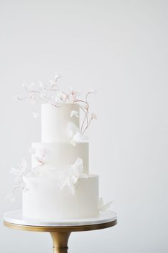 a three tiered white wedding cake with flowers on the top and bottom, sitting on a gold pedestal