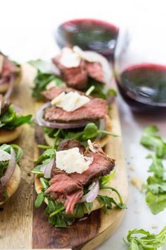 small sandwiches with meat, cheese and lettuce on wooden cutting board next to glass of red wine