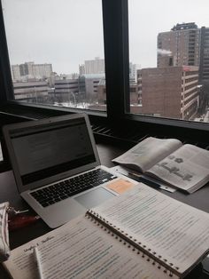 an open book sitting next to a laptop computer on top of a desk in front of a window