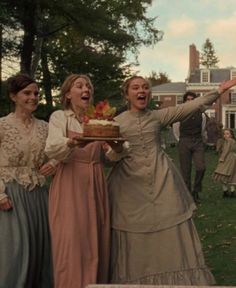 three women dressed in period clothing are holding a cake and smiling at the camera while others look on