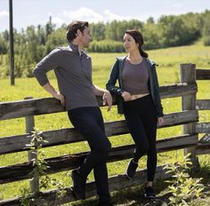 a man standing next to a woman on top of a wooden bench in front of a field