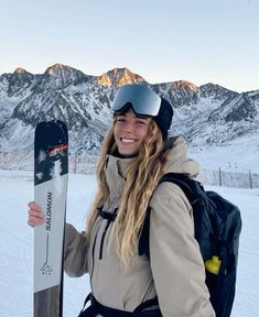 a woman holding a snowboard on top of a snow covered slope with mountains in the background