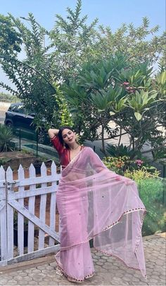 a woman in a pink sari standing next to a white fence with trees and bushes behind her