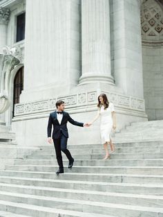 a man and woman holding hands while walking up some steps in front of a building