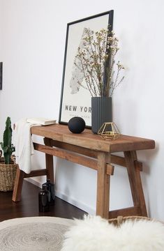 a wooden table with a vase and some plants on it next to a white wall