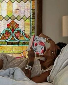 a woman laying in bed reading a book with a stained glass window behind her on the wall