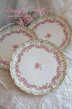 three plates with pink flowers on them sitting on a lace tablecloth next to a feather