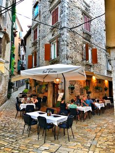 people are sitting at tables under an umbrella in front of a stone building with red shutters