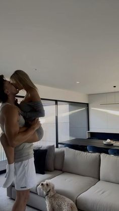 a woman holding a baby while standing next to a dog on the floor in front of a couch