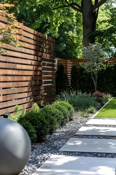 Modern garden with a wooden fence, green shrubs, a small tree, and a pathway with large rectangular stones. A gray spherical ornament sits by the fence. Design Garden Ideas, Living Room Loft