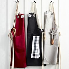 three aprons hanging from hooks on a white wall with red and black kitchen accessories