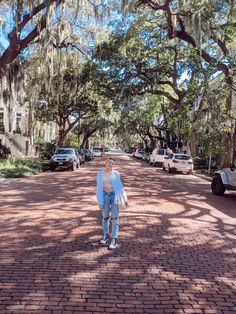 a woman walking down a brick road with cars parked on both sides and trees lining the street