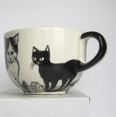 a black and white coffee cup with two cats painted on the side, sitting on top of a box