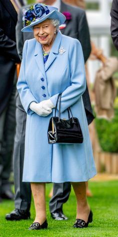 an older woman in a blue coat and hat holding a black purse while standing on grass