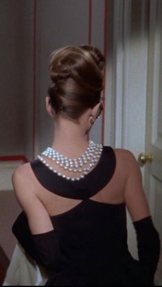 a woman in a black and white dress with pearls on her neck looking at herself in the mirror