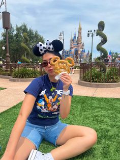 a woman sitting on the grass eating a pretzel in front of mickey mouse ears