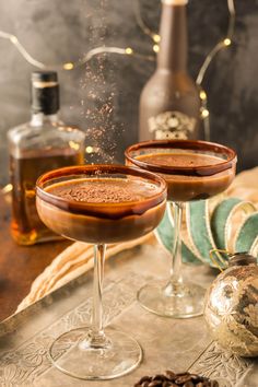 two glasses filled with liquid sitting on top of a table next to bottles and pine cones