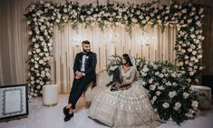 a bride and groom sitting on a couch in front of a flower covered wall at their wedding