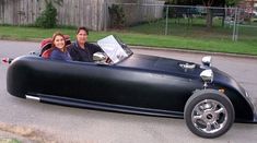 a man and woman sitting in an old fashioned car on the side of the road