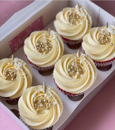 cupcakes with white frosting and gold decorations in a box on a pink surface