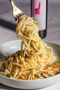 a fork full of spaghetti with parmesan cheese and herbs on it in a white bowl next to a bottle of wine