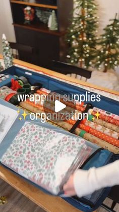 an open suitcase filled with wrapping supplies on top of a wooden table next to a christmas tree