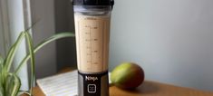 a blender sitting on top of a wooden table next to an apple and plant