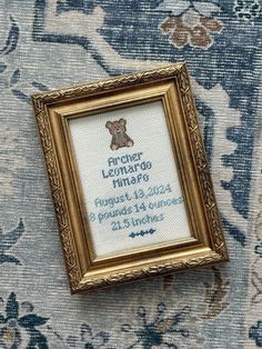 a framed cross - stitch teddy bear in a gold frame on a blue floral background