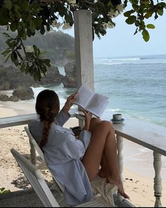 a woman sitting in a chair reading a book on the beach next to the ocean