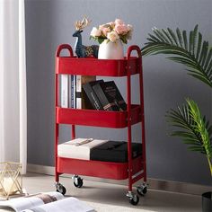 a red rolling cart with books and flowers on it next to a plant in a room