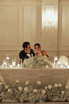 a bride and groom sitting at a table with candles on it, surrounded by flowers