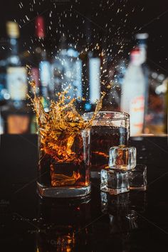 two glasses filled with liquid on top of a table next to liquor bottles and ice cubes