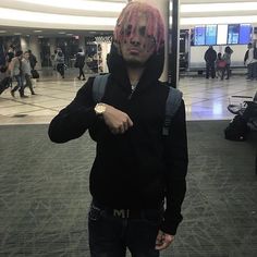 a man with pink hair wearing a black hoodie pointing at the camera in an airport