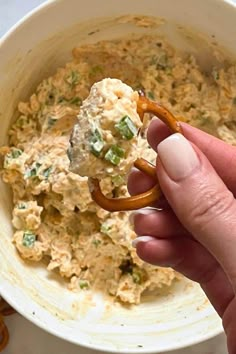 a hand holding a pretzel in front of a bowl of dip and crackers