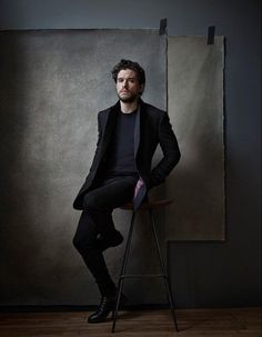 a man sitting on top of a chair in front of a gray wall with a painting behind him