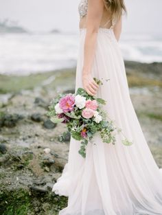 a woman in a white dress holding a bouquet on top of a rocky cliff near the ocean