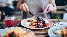 a woman is cutting into some pancakes with fruit on top and whipped cream on the side