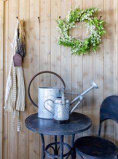 an old fashioned watering can sits on a table next to two chairs and a wreath