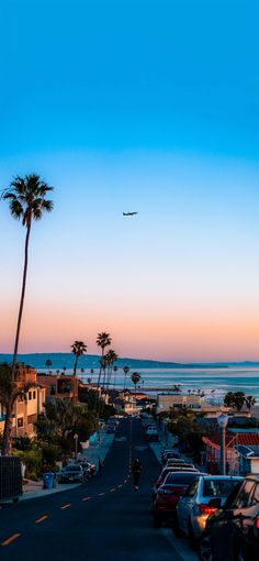 an airplane is flying over the beach and palm trees at sunset, with cars parked on the street below