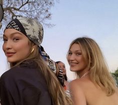 two beautiful women standing next to each other in front of a tree and one is wearing a bandana