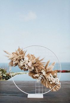 an arrangement of dried flowers and feathers on a circular stand in front of the ocean