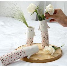 two white roses are in vases on a wooden tray and someone is holding one