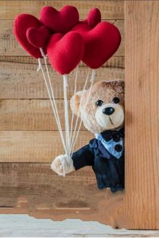 a stuffed teddy bear holding two red heart shaped balloons in it's mouth while hanging from the side of a wooden wall