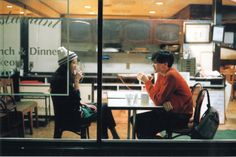 two people sitting at a table in front of a glass window talking on their cell phones