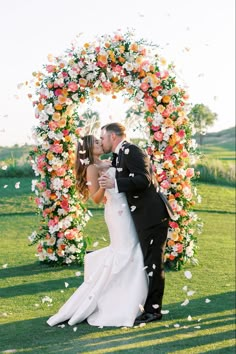 a bride and groom kissing in front of a floral arch with petals flying around them