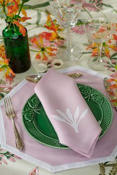 a place setting with pink napkins, green plates and silverware on a floral table cloth