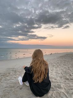 a woman sitting on top of a sandy beach next to the ocean with birds flying overhead