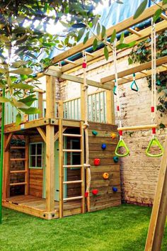 a wooden play structure with swings and climbing bars in the grass next to a brick wall